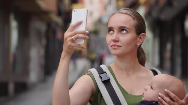 Mama stoi na ulicy z dzieckiem w chuście i strzela wideo do sieci społecznościowych, o jego podróży do telefonu komórkowego — Wideo stockowe