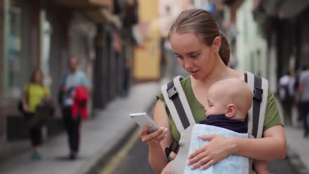 Mamma står på gatan med ett barn i en sele och tittar på skärmen mobiltelefon — Stockvideo