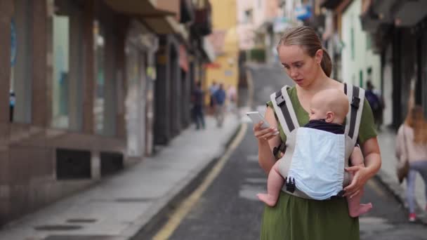 Mom is standing on the street with a baby in a sling and looks at the mobile phone screen and looks through the photos — Stock Video