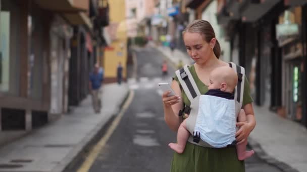 Mãe fica na rua com uma criança em uma funda e olha para a tela do telefone celular e se comunica em redes sociais — Vídeo de Stock