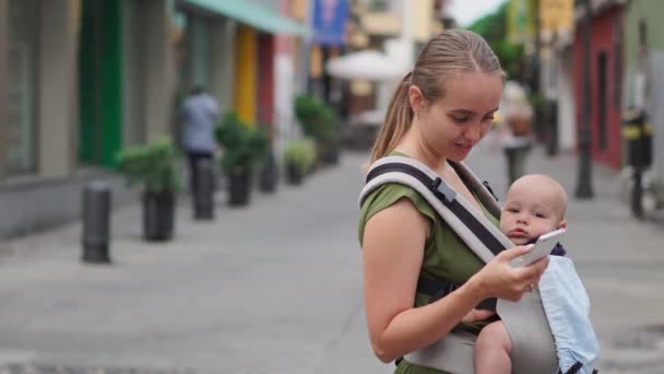 Mutter steht mit Baby im Tragetuch auf der Straße und schaut auf den Handybildschirm und drückt mit dem Finger auf den Bildschirm — Stockvideo