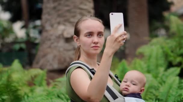 Мама с ребенком в стропе держит в руках смартфон и общается через видеосвязь с друзьями — стоковое видео