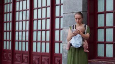 Genç bir anne gülümseyen kucağında, bir bebek tutan ve fon karşı sokakta yürürken onu smartphone bir fotoğrafı çeker. Annelik ve bloglama.