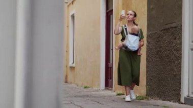 Genç bir anne gülümseyen kucağında, bir bebek tutan ve fon karşı sokakta yürürken onu smartphone bir fotoğrafı çeker. Annelik ve bloglama.