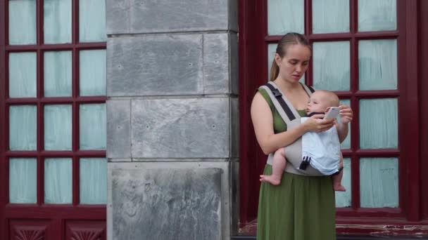 Una joven madre sostiene a un bebé en sus brazos, sonriendo y toma una foto en su teléfono inteligente mientras camina por la calle contra el telón de fondo. Maternidad y blogueo . — Vídeos de Stock