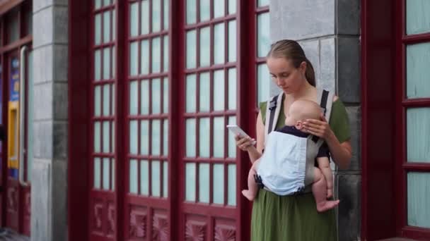 Una joven madre sostiene a un bebé en sus brazos, sonriendo y toma una foto en su teléfono inteligente mientras camina por la calle contra el telón de fondo. Maternidad y blogueo . — Vídeos de Stock