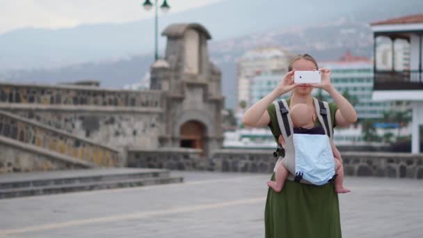 Una giovane madre tiene un bambino tra le braccia, sorride e scatta una foto sul suo smartphone mentre cammina per strada sullo sfondo. Maternità e blogging . — Video Stock