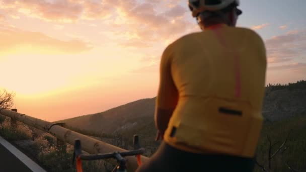 Чоловік-велосипедист у жовтій футболці в горах спостерігає за заходом сонця. Відпочинок після тренування, залізна людина. Вид ззаду — стокове відео