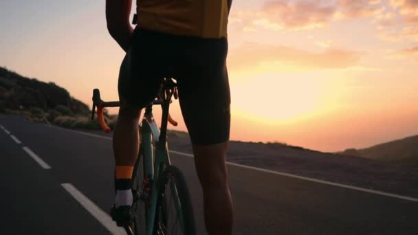 Hombre ciclista en una camiseta amarilla en las montañas observando la puesta de sol. Descansando después de un entrenamiento, hombre de hierro. La vista desde atrás — Vídeos de Stock