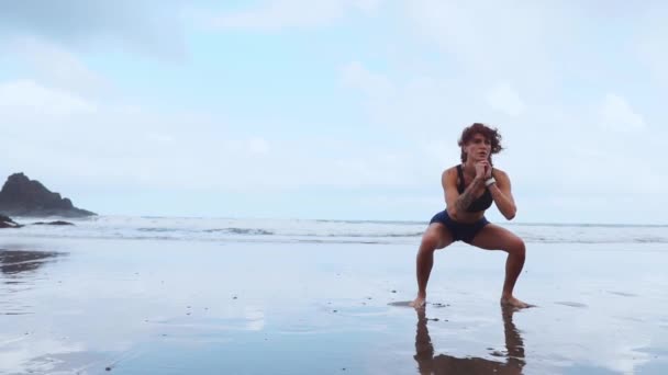 Ajuste piernas de entrenamiento de mujer con ejercicios de salto de entrenamiento de hiit sentadillas ejercicios. Entrenamiento de fitness haciendo ejercicio cardiovascular en la playa de arena blanca del océano de verano haciendo saltos explosivos y eructos para activar los glúteos. — Vídeos de Stock