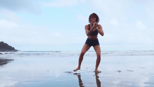 Ajuste piernas de entrenamiento de mujer con ejercicios de salto de entrenamiento de hiit sentadillas ejercicios. Entrenamiento de fitness haciendo ejercicio cardiovascular en la playa de arena blanca del océano de verano haciendo saltos explosivos y eructos para activar los glúteos. — Vídeo de stock