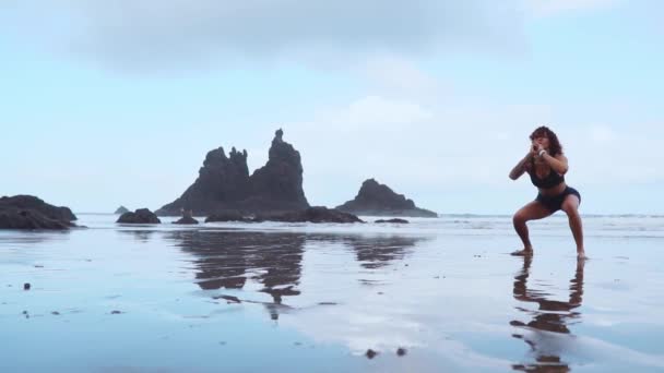 Ragazza salta fuori dopo un accovacciato in allenamento sulla spiaggia con sabbia nera vicino all'oceano Atlantico sullo sfondo delle montagne — Video Stock