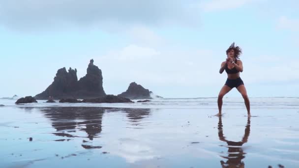 Dívka skáče po dřepu v tréninku na pláži s černým pískem poblíž Atlantský oceán na pozadí hor — Stock video