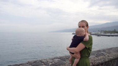 Okyanus ve dalgaların çarpıcı görünümü bakarak elleri üzerinde bir bebek ile genç bir anne vuruyor. Çocuk gemi uzak gösterir.