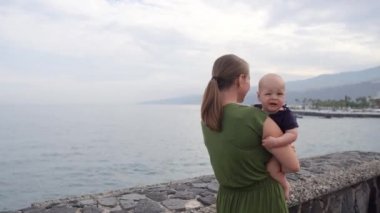 Okyanus ve dalgaların çarpıcı görünümü bakarak elleri üzerinde bir bebek ile genç bir anne vuruyor. Çocuk gemi uzak gösterir.