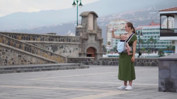 Μια νεαρή γυναίκα με ένα μωρό σε μια σφεντόνα ταξιδεύει στην Ευρώπη. Στέκεται στην πλατεία κοντά στον ωκεανό παίρνει για τα αξιοθέατα, τηλέφωνο και είναι μεταδίδονται σε απευθείας σύνδεση για το blog σας — Αρχείο Βίντεο