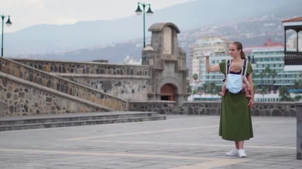 Młoda kobieta z dzieckiem w chuście przemieszcza się do Europy. Stojąc na placu, w pobliżu oceanu na telefon atrakcji i jest transmisję online na swoim blogu — Wideo stockowe