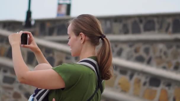 Молодая женщина с ребенком в стропе едет в Европу. Стоя на площади рядом с океаном берет на телефон достопримечательностей и транслируется онлайн для вашего блога — стоковое видео