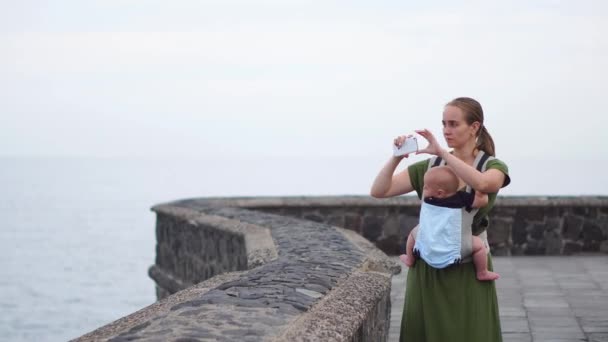 Una madre joven con un bebé dispara en sus manos mirando al océano y dispara una vista impresionante de las olas en su teléfono inteligente — Vídeos de Stock