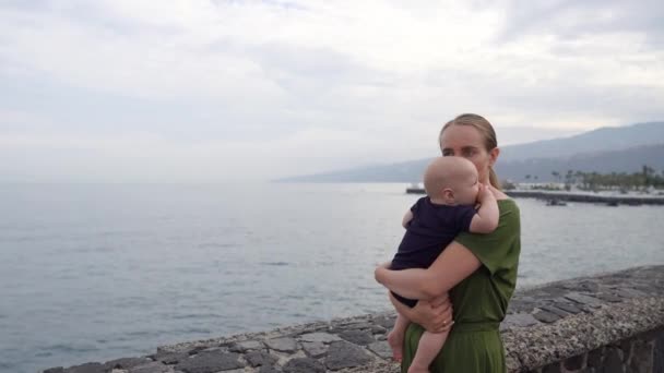 En ung mamma med en baby skjuter på händerna tittar på havet och den fantastiska utsikten av vågorna. Visar barnet fartyg i fjärran. — Stockvideo