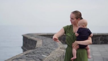 Okyanus ve dalgaların çarpıcı görünümü bakarak elleri üzerinde bir bebek ile genç bir anne vuruyor. Çocuk gemi uzak gösterir.