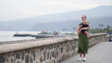 Genç Anne oğlu bebeği ile okyanusu Europe bakar, gülümser ve dalgalar antik meydanında yakın sahildeki yürür