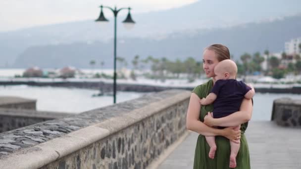 Ung mamma promenader med sin son baby vid vattnet nära havet på det gamla torget i Europa ser på vågorna och leenden — Stockvideo