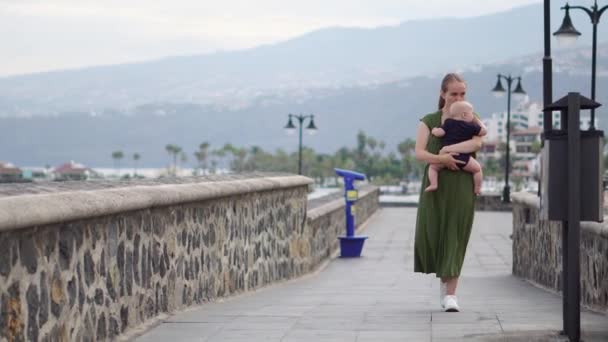 年轻的母亲散步与她的儿子婴孩在海滨附近的海洋在欧洲的古老广场看着海浪和微笑 — 图库视频影像