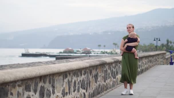 Junge mutter spaziert mit ihrem sohn baby am ufer in der nähe des meeres auf dem antiken platz von europa blickt auf die wellen und lächelt — Stockvideo