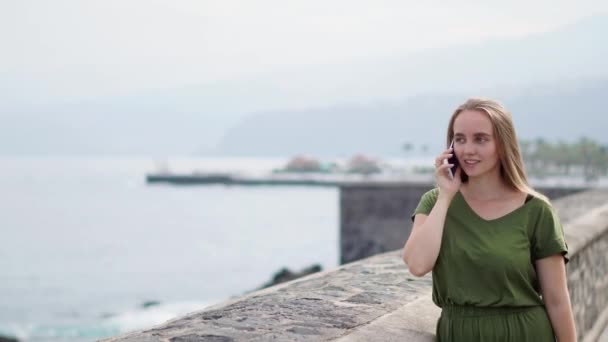 Hermosa chica en un vestido verde hablando por teléfono de pie en el paseo marítimo cerca del océano y sonriendo — Vídeos de Stock
