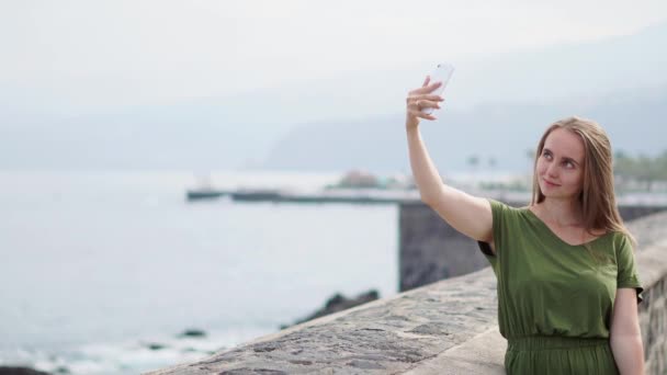 一个穿着绿色衣服的漂亮女孩在靠近海洋的海滨的一个手机上自拍, 微笑着 — 图库视频影像