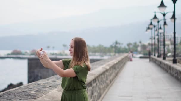 Красива дівчина в зеленій сукні знімає відео для блогу на мобільному телефоні, що стоїть на набережній біля океану і посміхається — стокове відео