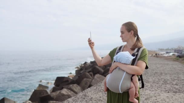 En ung kvinna i en klänning med en baby tar av havet på en mobiltelefon som går längs Atlantkusten i Kanarieöarna. Resa i Europa — Stockvideo