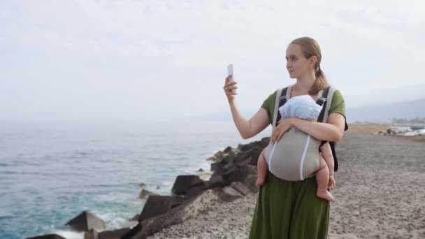 Μια νεαρή γυναίκα σε ένα φόρεμα με ένα μωρό παίρνει μακριά στον ωκεανό σε ένα κινητό τηλέφωνο, περπατώντας κατά μήκος της ατλαντικής ακτής στα Κανάρια νησιά. Ταξίδια στην Ευρώπη — Αρχείο Βίντεο