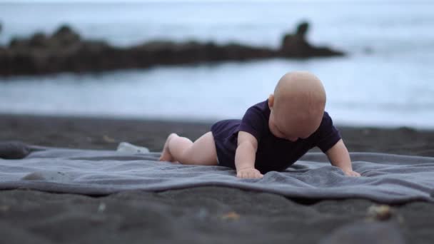 The baby lies on his stomach on the black sand near the ocean and laughs looking at the camera — Stock Video