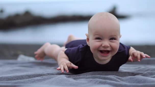 Le bébé s'allonge sur le ventre sur le sable noir près de l'océan et rit en regardant la caméra — Video