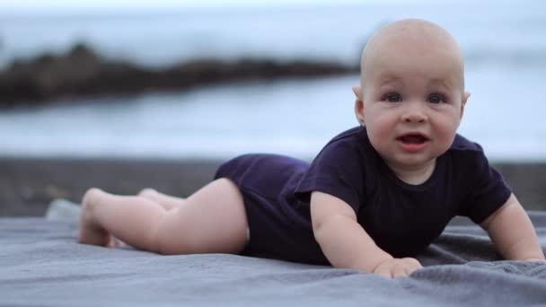 Дитина лежить на животі на чорному піску біля океану і сміється дивитися на камеру — стокове відео