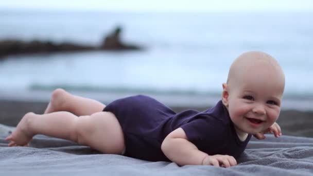 Il bambino giace sul suo stomaco sulla sabbia nera vicino all'oceano e ride guardando la macchina fotografica — Video Stock