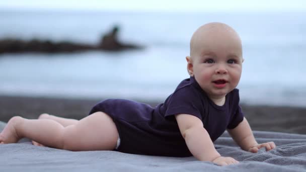 Dítě leží na břiše na černém písku nedaleko oceánu a směje se, při pohledu na fotoaparát
