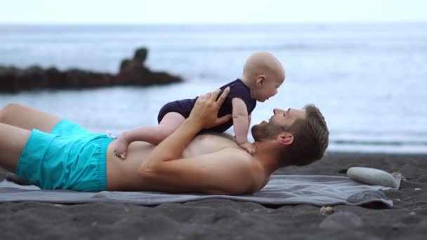 Padre jugando con su hijo playa bebé en la playa de arena negra en el fondo del océano Atlántico. Tomar de la mano y reír — Vídeo de stock
