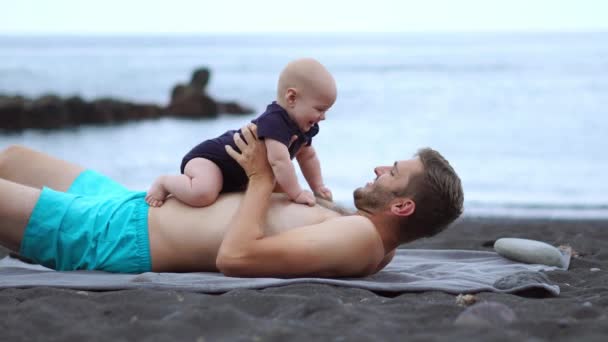 Pai brincando com seu filho praia do bebê na praia de areia preta no fundo do oceano Atlântico. De mãos dadas e a rir. Simular voo — Vídeo de Stock
