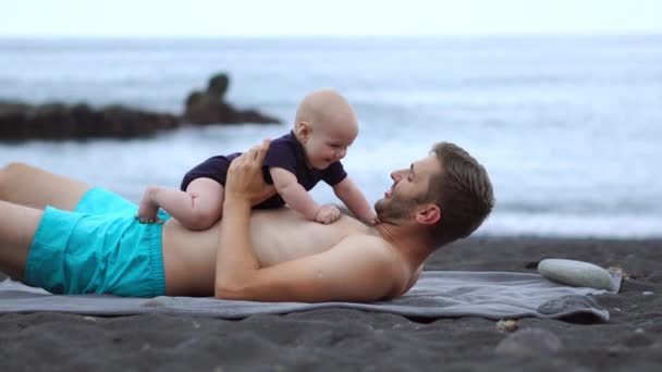 Padre jugando con su hijo playa bebé en la playa de arena negra en el fondo del océano Atlántico. Tomando de la mano y riendo. Para simular el vuelo y abrazos — Vídeo de stock