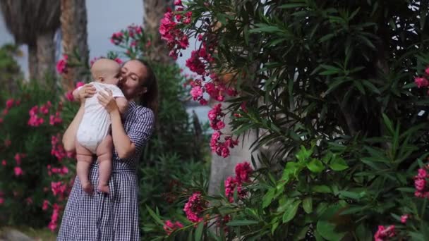 Madre a hijo juega un abrazo y besa al bebé alrededor de las palmeras con flores en el resort. Familia feliz — Vídeo de stock