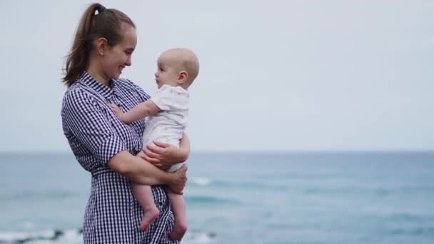 美丽的母亲的肖像亲吻女儿在海边和海滩的背景。快乐的家庭在假期。旅行。妈妈抱着孩子在日落时分。白人女性与婴孩在海洋外面。孕产妇护理. — 图库视频影像