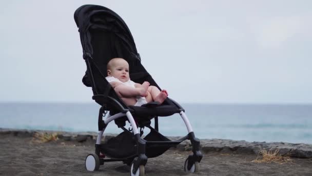Дитина в колясці на піщаному пляжі, мирно дивлячись на нескінченність і морські хвилі з великим інтересом, насолоджуючись пізнього літнього вечора біля моря. Сімейний відпочинок, подорожі з дітьми концепція . — стокове відео
