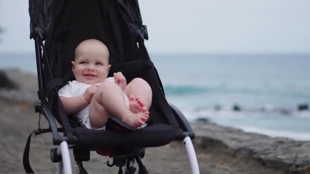 Baby in een kinderwagen op een zandstrand, rustig op zoek naar oneindigheid en zee golven met veel belangstelling, genieten van de nazomer avond door de zee. Familievakantie, reizen met kinderen concept. — Stockvideo