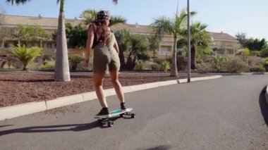 Ağır çekimde longboard palmiye ağaçlarının yakınındaki yolda genç kız rides