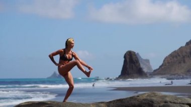 Yoga duran siyah bir kum Plajı üzerinde okyanus tarafından bir kayaya yapıyor tek ayak üzerinde duran Dengeleme bikinili genç kız