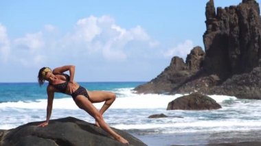 Kadın fitness volkanik siyah kum lav dağlarda bir kayanın üzerine oturmuş bir mayo ile sahilde yapıyor. Sağlıklı yaşam tarzı ve eğlence adalarda kavramı