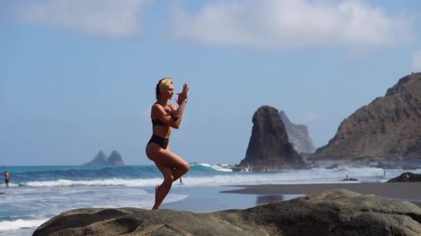 Ung flicka i bikini balansera stående på ett ben gör yoga står på en klippa vid havet på en svart sandstrand — Stockvideo